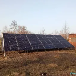 Instalacja fotowoltaiczna na gruncie, widok z boku na panele słoneczne zamontowane na metalowej konstrukcji, w tle budynek gospodarczy z cegły.