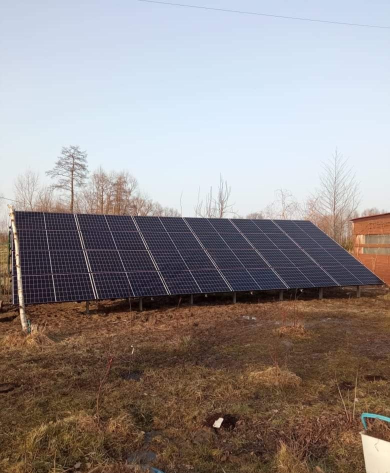 Instalacja fotowoltaiczna na gruncie, widok z boku na panele słoneczne zamontowane na metalowej konstrukcji, w tle budynek gospodarczy z cegły.