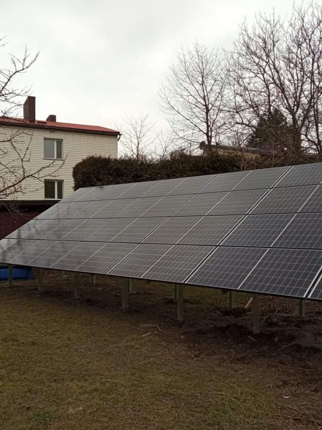 Instalacja paneli słonecznych na gruncie, widoczne metalowe podpory, w tle dom z czerwonym dachem i drzewa bez liści pod pochmurnym niebem.