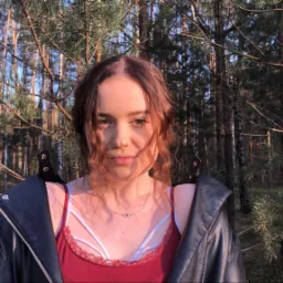 Portret młodej kobiety w bordowej koszulce i czarnej skórzanej kurtce na tle leśnym w promieniach zachodzącego słońca, z widocznym delikatnym naszyjnikiem.