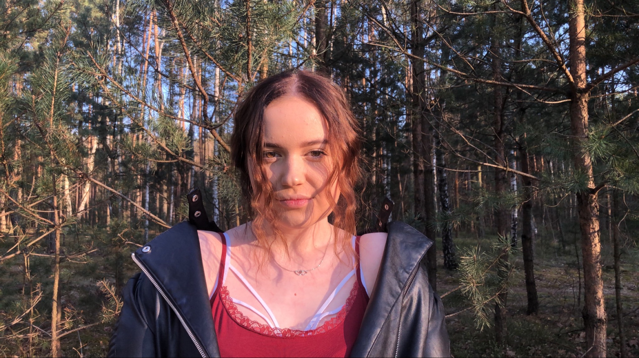 Portret młodej kobiety w bordowej koszulce i czarnej skórzanej kurtce na tle leśnym w promieniach zachodzącego słońca, z widocznym delikatnym naszyjnikiem.