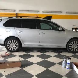 Srebrny samochód kombi marki Volkswagen z przyciemnionymi szybami bocznymi i tylną, stojący w warsztacie z podłogą w szachownicę, widoczne butelki ze spryskiwaczami i kartonowe pudełko.