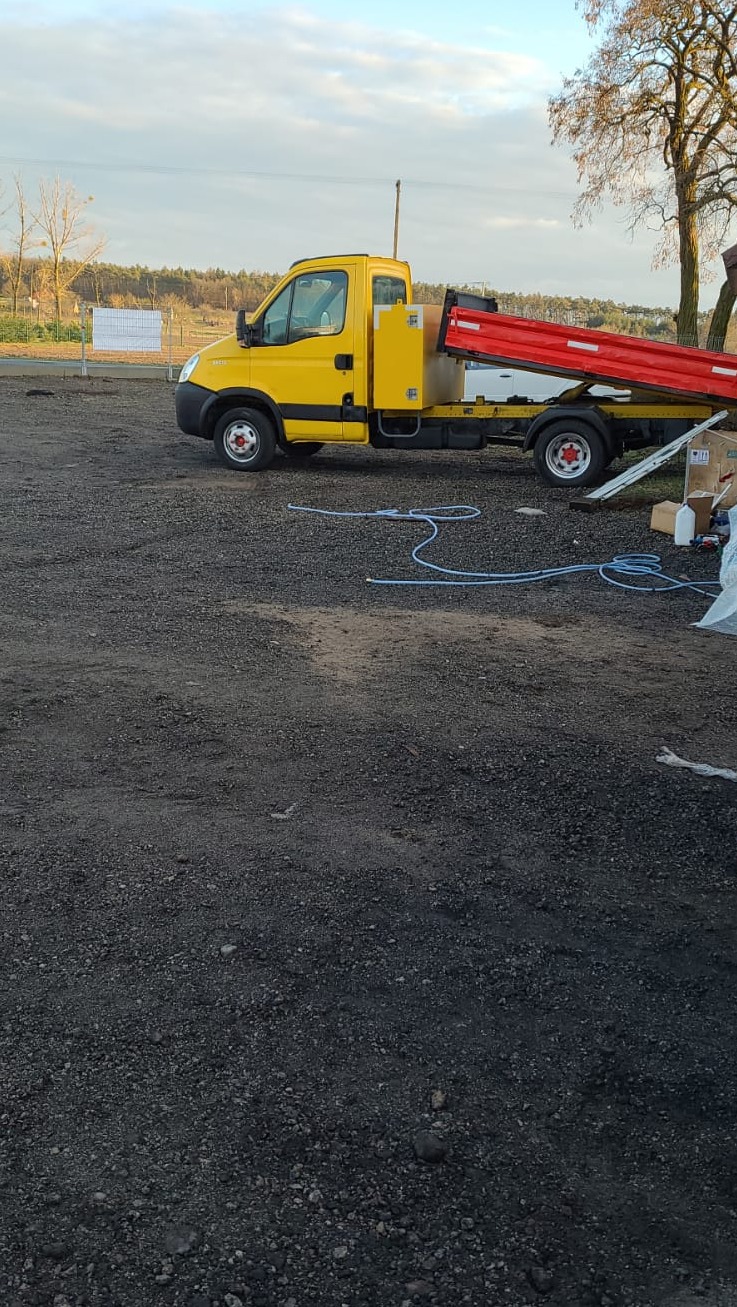 Żółta wywrotka z podniesioną, czerwoną skrzynią ładunkową stoi na żwirowym placu; w tle drzewa i błękitne niebo z chmurami.