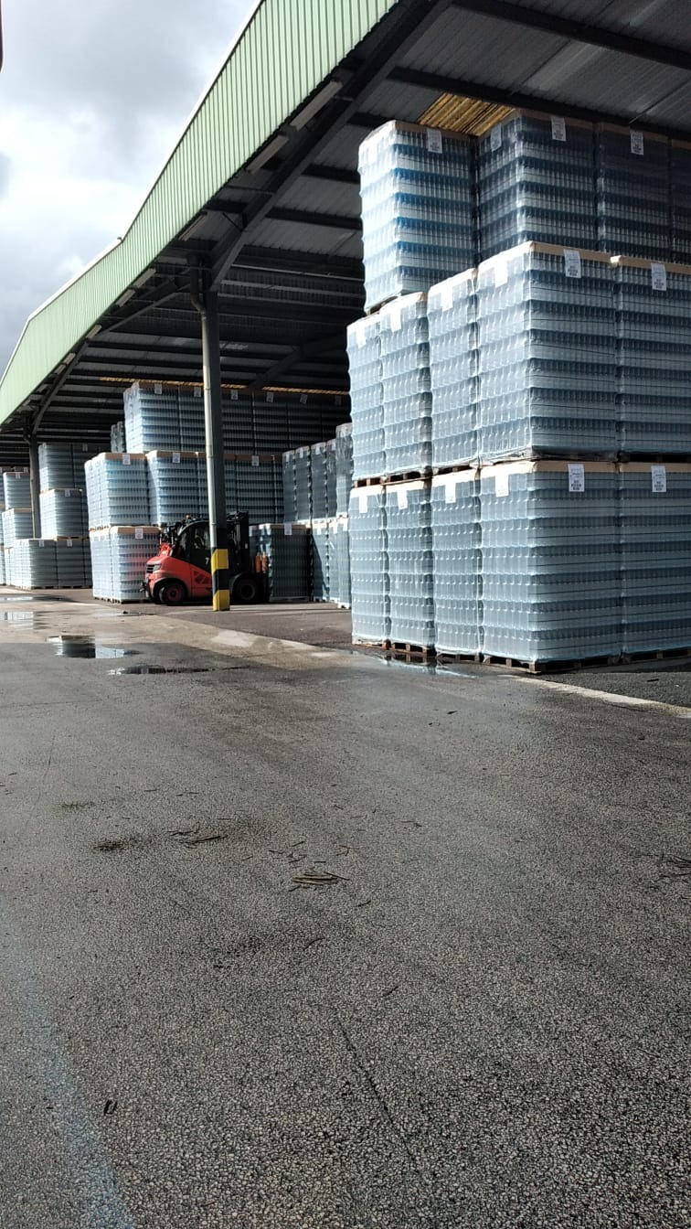 Magazyn zadaszony z paletami ułożonymi w stosy, owiniętymi folią, z wózkiem widłowym w tle, mokra nawierzchnia.