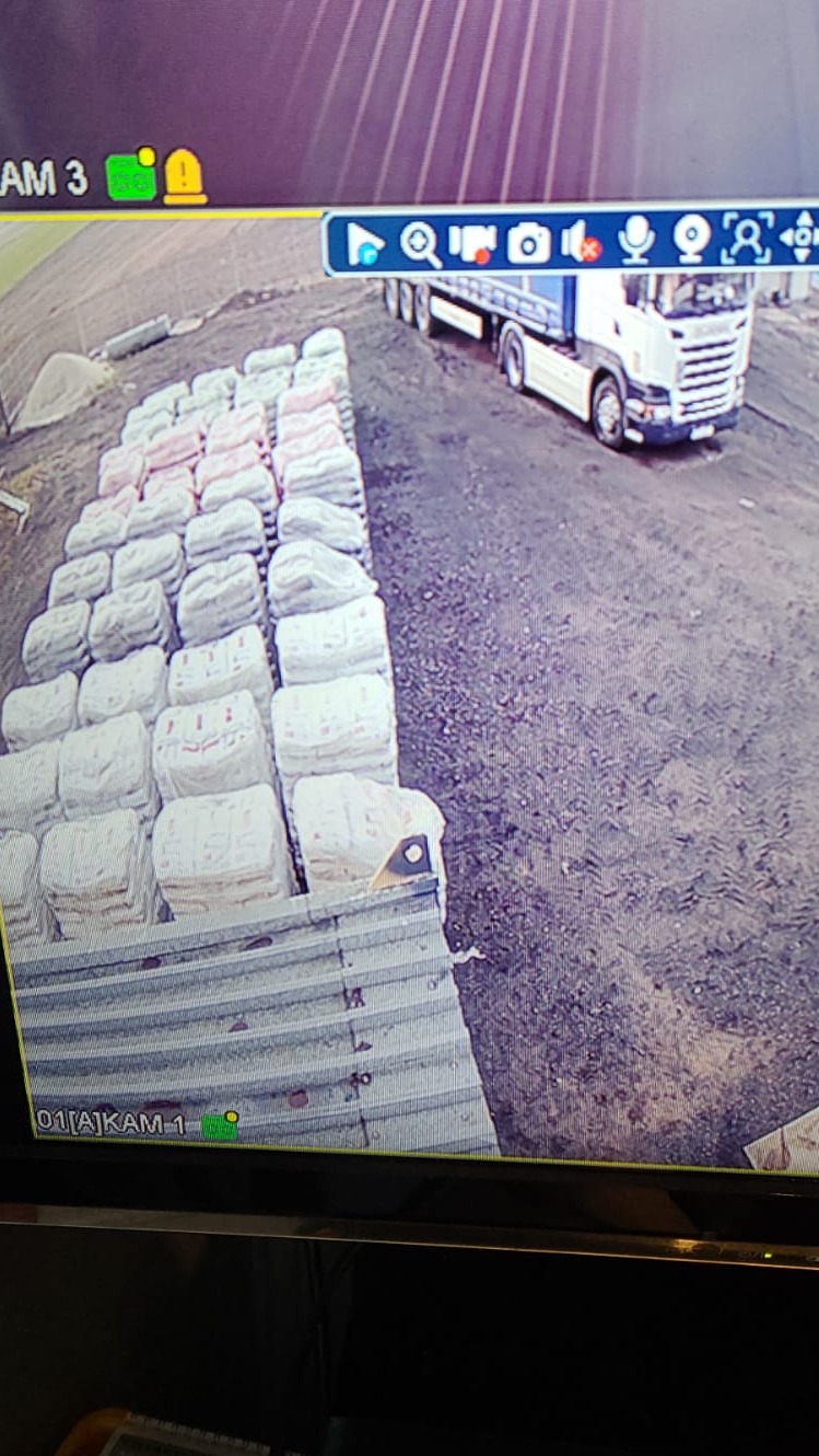 Widok z kamery przemysłowej na transport worków z opałem na naczepie ciężarówki, ustawionych w rzędach na placu składowym.