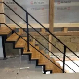 Nowoczesne schody wewnętrzne z drewnianymi stopniami i czarną metalową balustradą, widoczne na tle surowych ścian w trakcie remontu.