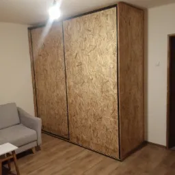 Duża szafa przesuwna z płyty OSB z czarnym obramowaniem w jasnym pokoju z drewnianą podłogą, widoczny fragment szarej sofy i mały stolik.