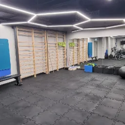 Wnętrze siłowni z drabinkami gimnastycznymi, matami do ćwiczeń, piłkami i bieżniami w tle, oświetlone geometrycznymi lampami sufitowymi.