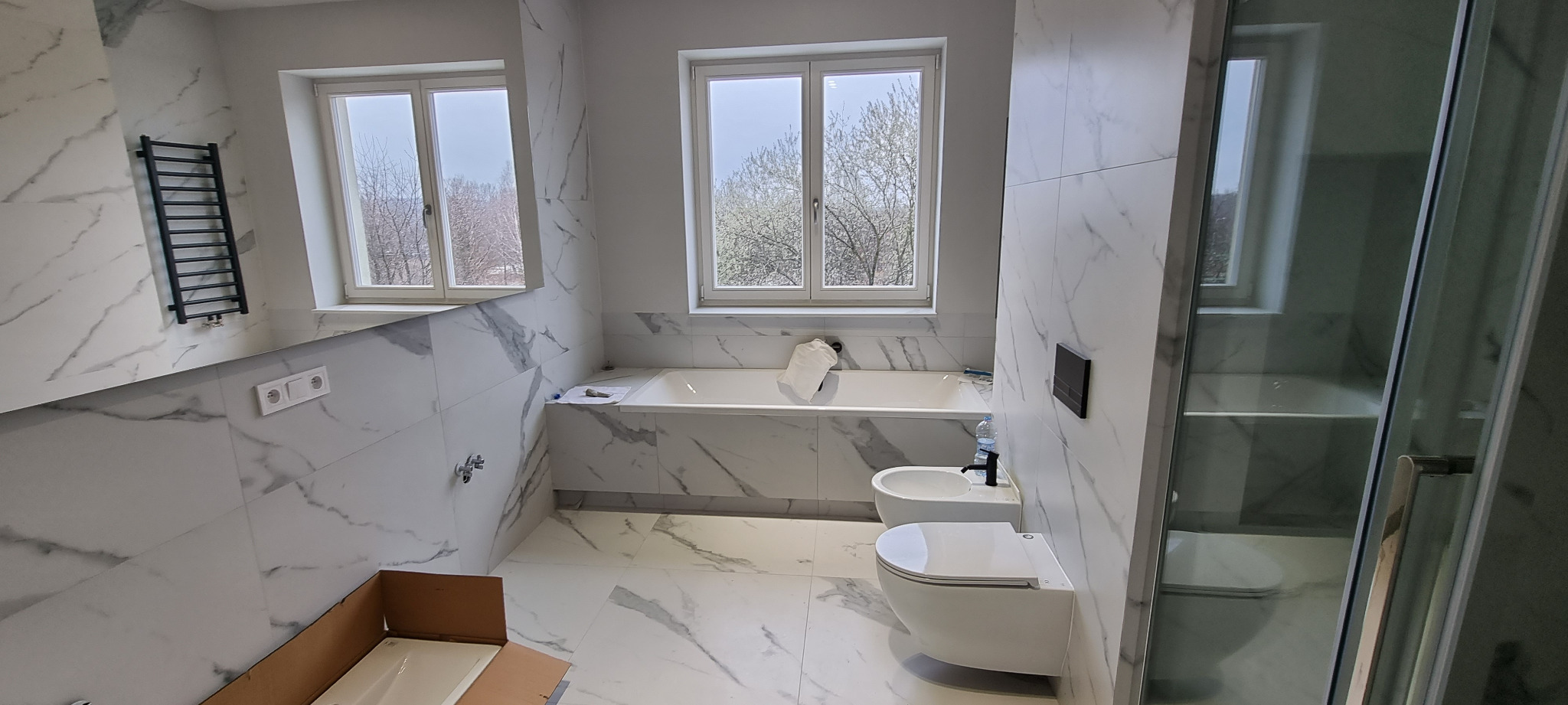 Nowoczesna łazienka z białymi płytkami imitującymi marmur, wisząca toaleta i bidet, wanna obudowana płytkami, grzejnik drabinkowy oraz kabina prysznicowa ze szklanymi drzwiami.