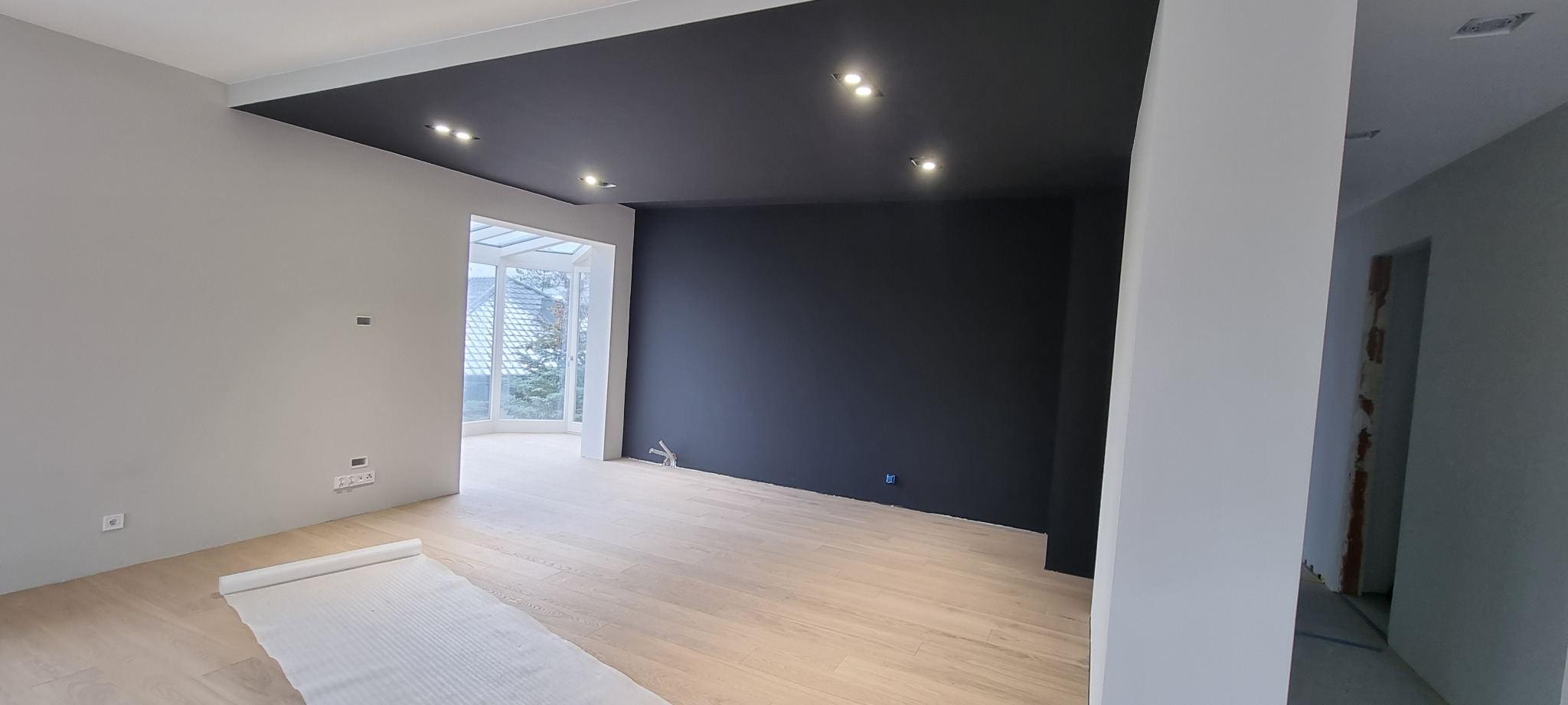 Nowoczesne wnętrze po remoncie: Jasne panele podłogowe, szara ściana, czarny sufit z oświetleniem punktowym oraz fragment czarnej ściany. Podłoga zabezpieczona folią malarską.