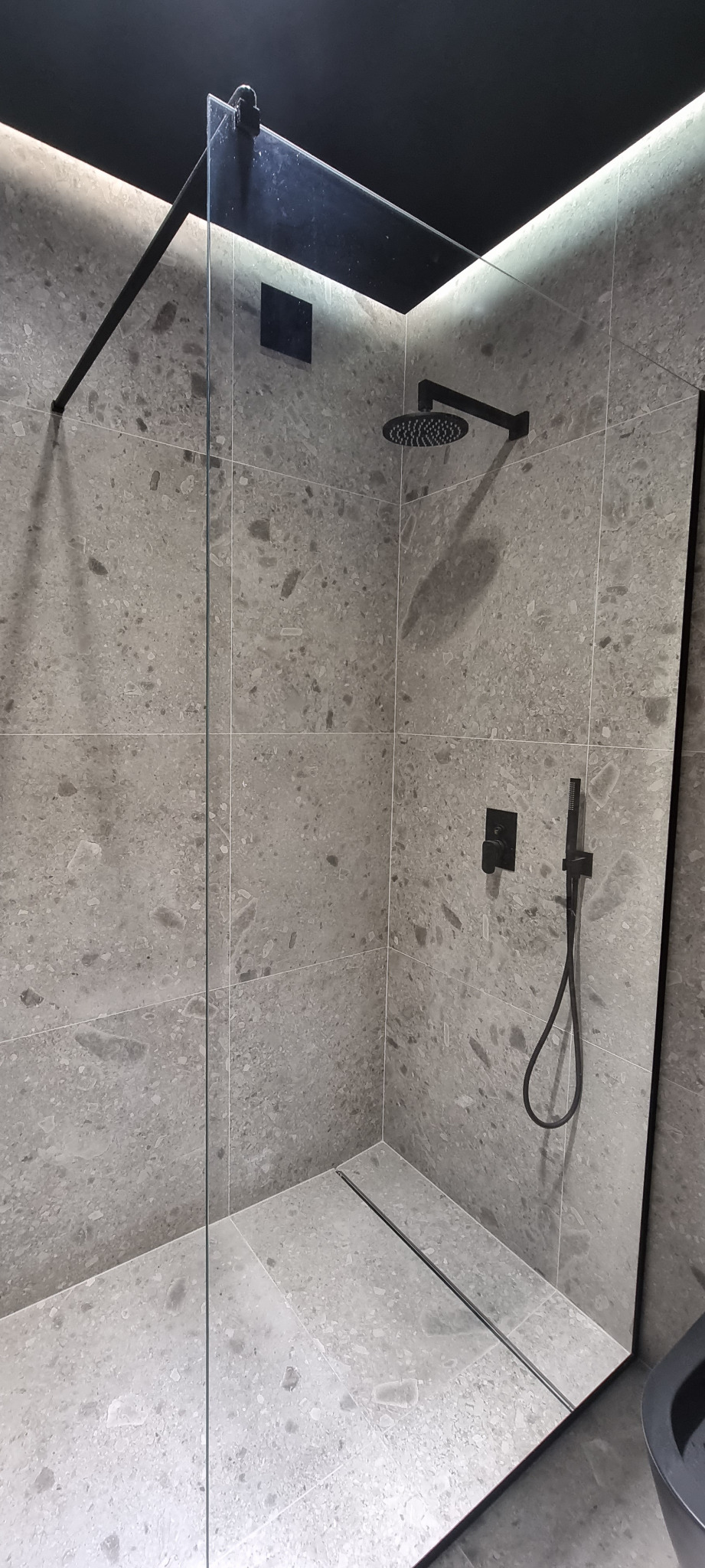 Nowoczesna kabina prysznicowa walk-in z szarą, kamienną okładziną, czarną armaturą i odpływem liniowym, oświetlona subtelnym światłem LED.