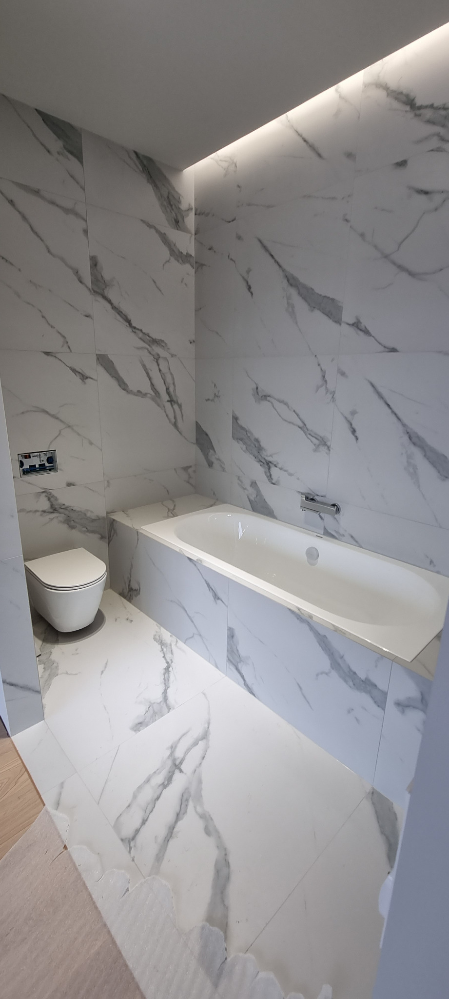 Nowoczesna łazienka z białymi płytkami imitującymi marmur, podświetlonym sufitem, wiszącą toaletą i wanną z zabudową.