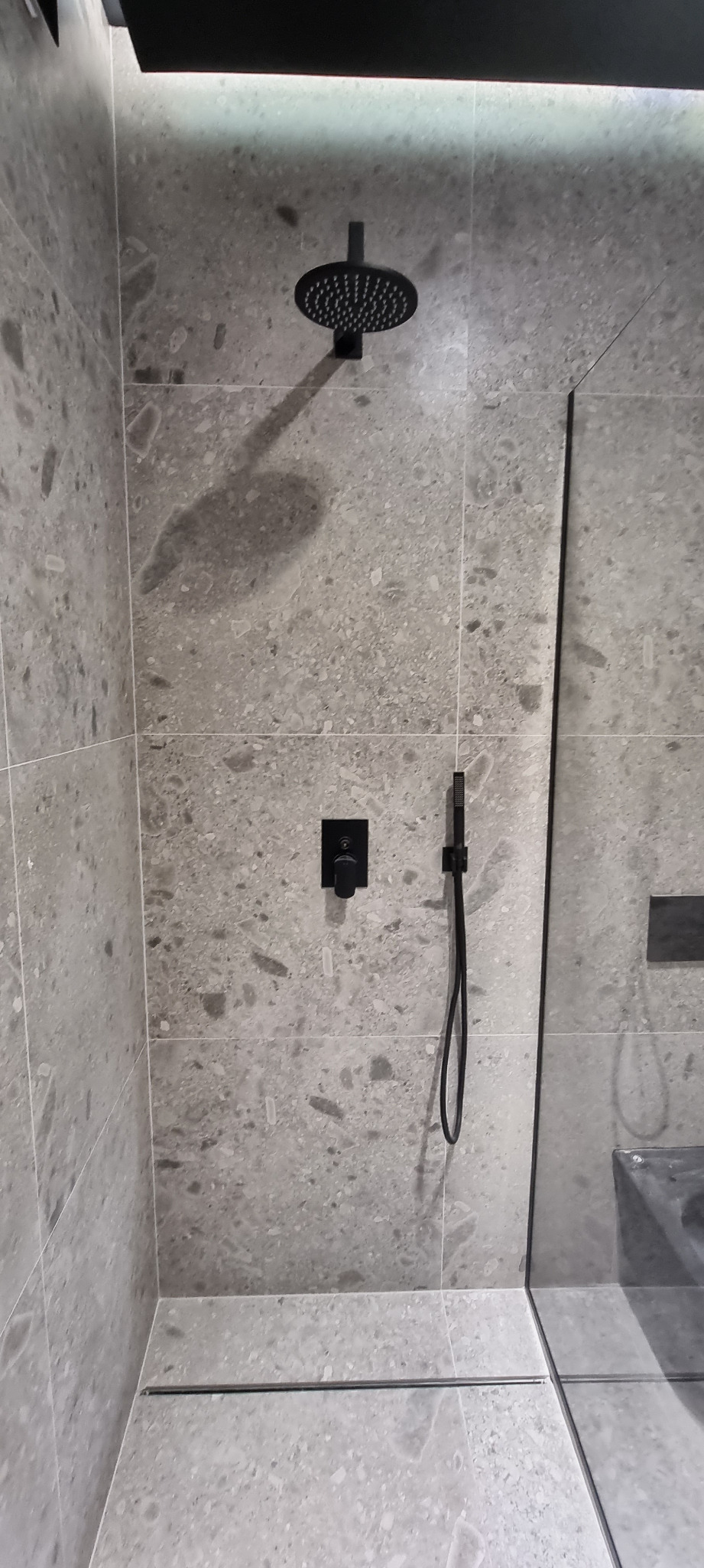 Nowoczesna kabina prysznicowa z szarymi płytkami imitującymi kamień, czarną deszczownicą i armaturą, minimalistycznym odpływem liniowym i szklaną ścianką.