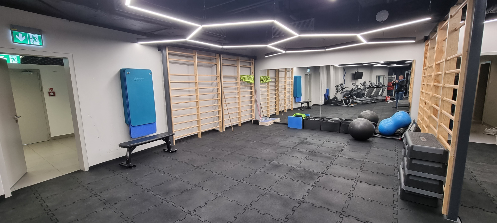 Wnętrze siłowni z drabinkami gimnastycznymi, matami do ćwiczeń, piłkami i bieżniami w tle, oświetlone geometrycznymi lampami sufitowymi.