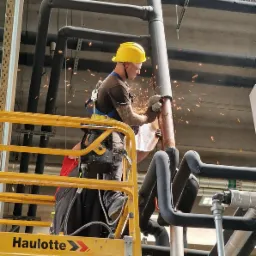 Pracownik w kasku i okularach ochronnych na podnośniku Haulotte szlifuje rurę instalacyjną, iskry lecą, widoczne liczne rury w izolacji.