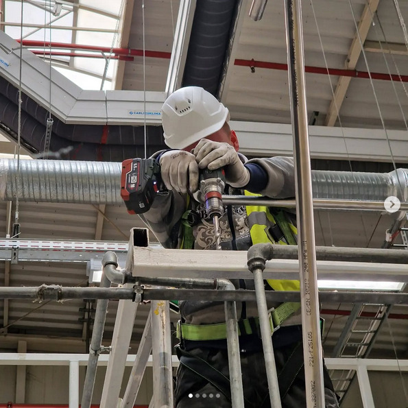 Pracownik w kasku i kamizelce odblaskowej wierci otwór w metalowej konstrukcji, otoczony rurami i elementami instalacji przemysłowej, widoczne przewody wentylacyjne pod sufitem.