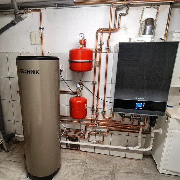 Kotłownia z kotłem Buderus, zasobnikiem Technix i instalacją z miedzianych rur, manometry na czerwonych zbiornikach, widoczne płytki na ścianach i podłodze