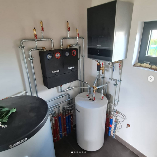 Złożona instalacja grzewcza Buderus z dwoma czarnymi modułami, białym zasobnikiem i kotłem wiszącym, liczne rury miedziane i stalowe, widok z boku, pomieszczenie techniczne.