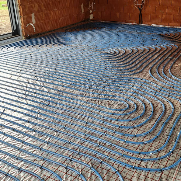 Niebieskie rury ogrzewania podłogowego ułożone spiralnie na srebrnej folii izolacyjnej i czerwonej siatce w surowym wnętrzu z ceglanymi ścianami.