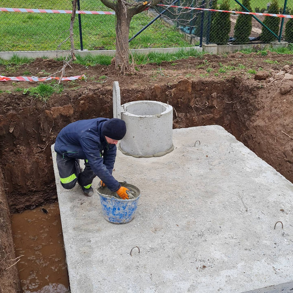 Pracownik w pomarańczowych rękawicach uszczelnia betonową płytę szamba z kręgami na wykopie ziemnym, widoczne narzędzia i woda w wykopie.