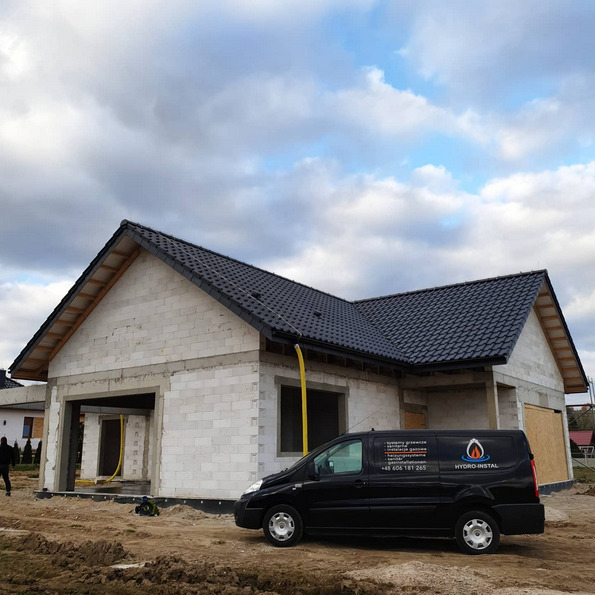 Surowy stan domu w budowie z podłączonym żółtym przewodem gazowym i zaparkowanym czarnym vanem firmy instalacyjnej na tle pochmurnego nieba.