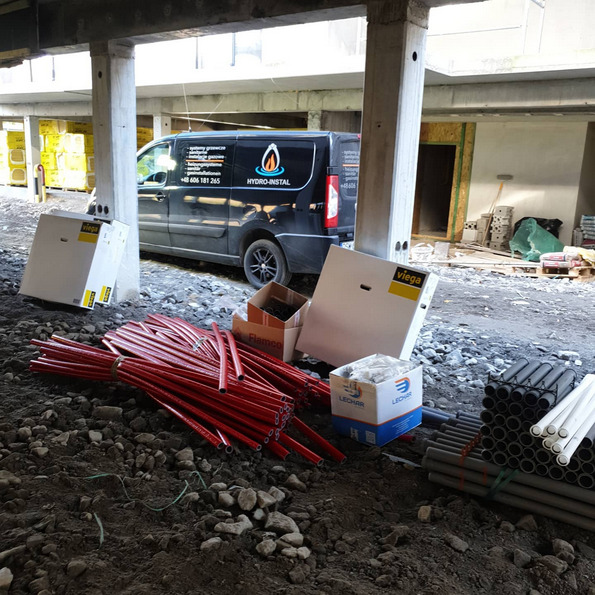 Miejsce budowy z ułożonymi rurami instalacyjnymi w kolorze czerwonym i szarym, kartonami Viega i Lechar, oraz zaparkowanym vanem firmy hydraulicznej Hydro-Instal w tle.