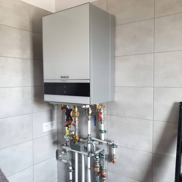Zamontowany gazowy piec centralnego ogrzewania marki Buderus z podłączeniem do instalacji grzewczej, widoczne rury i zawory, na tle jasnych płytek ceramicznych.