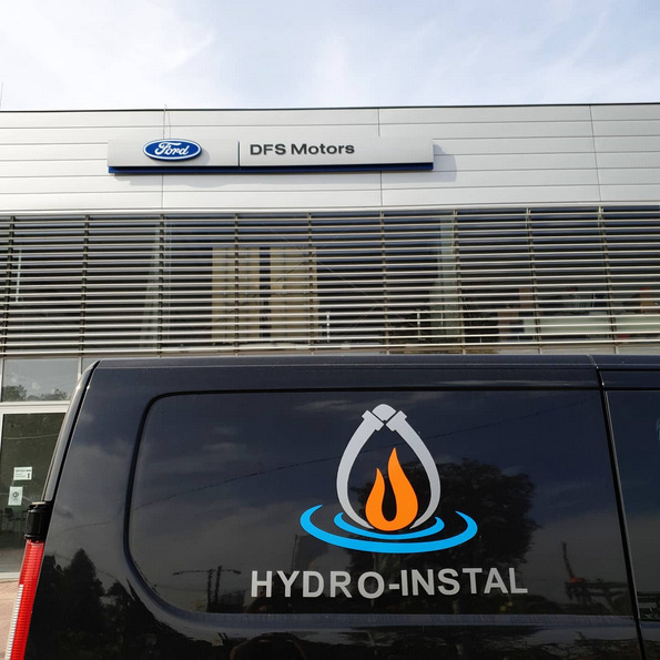 Czarny samochód dostawczy z logo firmy Hydro-Instal, przedstawiającym płomień w kropli wody otoczony rurami, zaparkowany przed budynkiem salonu Ford DFS Motors.