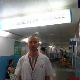 Lekarz w białym fartuchu z logo szpitala na tle korytarza kliniki medycyny wschodniej i zachodniej, z pacjentami i oznaczeniami w języku chińskim i angielskim.