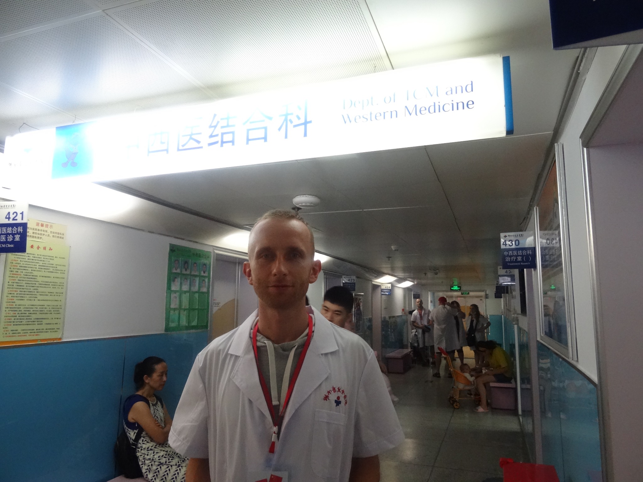 Lekarz w białym fartuchu z logo szpitala na tle korytarza kliniki medycyny wschodniej i zachodniej, z pacjentami i oznaczeniami w języku chińskim i angielskim.