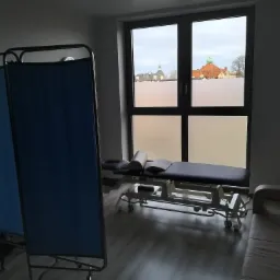 Gabinet fizjoterapii z kozetką i parawanem, widok na miasto przez okno z matową folią.