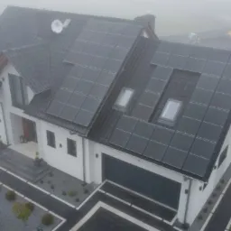 Dom jednorodzinny z zamontowaną instalacją fotowoltaiczną na dachu, widoczny w porannej mgle, z zagospodarowanym terenem wokół, w tle plac zabaw.