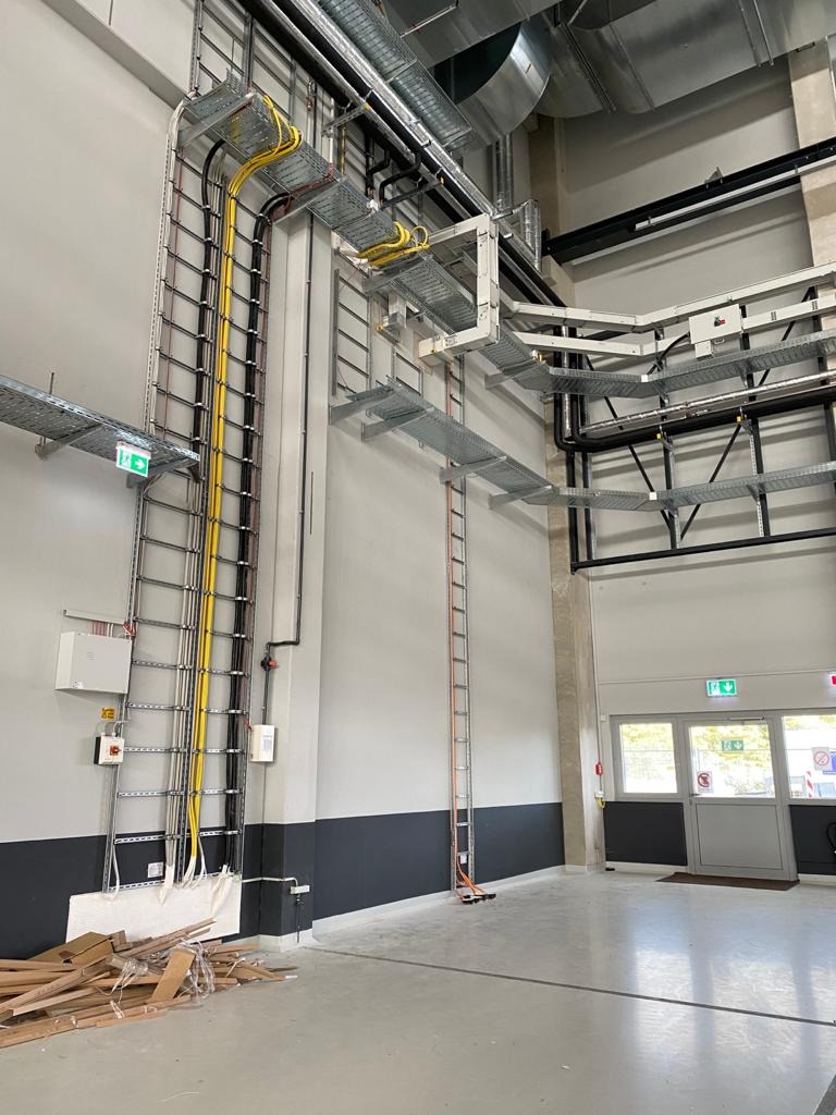 Wysokie pomieszczenie przemysłowe z szynami kablowymi pod sufitem i pionowymi prowadnicami kablowymi przy ścianie, żółte i czarne kable ułożone w korytach, skrzynka elektryczna i włączniki...