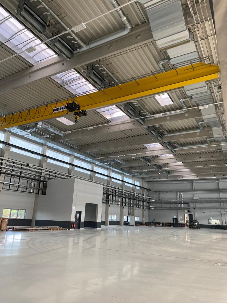 Wnętrze hali przemysłowej z widocznymi instalacjami pod sufitem, w tym żółty suwnicowy dźwig podwieszany z napisem NEWI, liczne rury wentylacyjne i oświetlenie rastrowe.