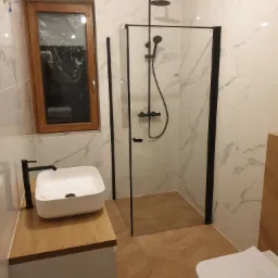 Nowoczesna łazienka po remoncie: kabina prysznicowa z czarnymi ramami, biała umywalka nablatowa z czarną armaturą, marmurowe płytki ścienne i drewnopodobna podłoga w jodełkę, okno z folią ochronną.