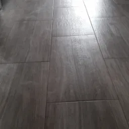 Ułożona podłoga z szarych, matowych płytek imitujących drewno, widok z perspektywy podłogi w kierunku rozświetlonego wejścia.