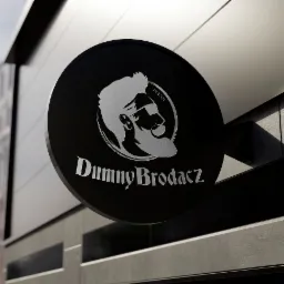 Elegancki, czarny szyld z logo DumnyBrodacz.pl w srebrnym kolorze, przedstawiającym profil brodatego mężczyzny w okularach, zamontowany na elewacji budynku.