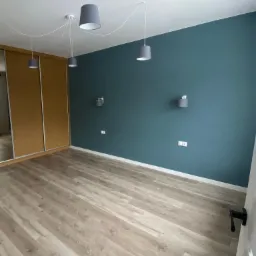 Pokój po remoncie: ściana w kolorze petrol, jasna podłoga z paneli, przesuwne drzwi szafy z lustrem, lampy wiszące z szarymi abażurami i kinkiety.