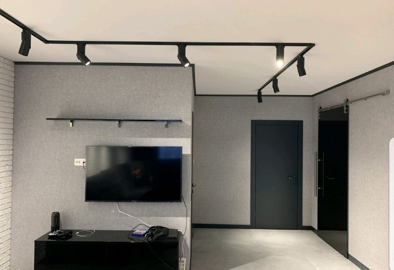 Szare wnętrze pokoju z oświetleniem szynowym, telewizorem na ścianie i przesuwnymi drzwiami, minimalistyczny design.