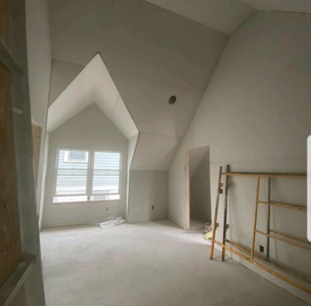 Poddasze w trakcie prac wykończeniowych: ściany pokryte szpachlą, widoczne łączenia płyt, drewniany ruszt na pierwszym planie, okno w szczycie dachu.