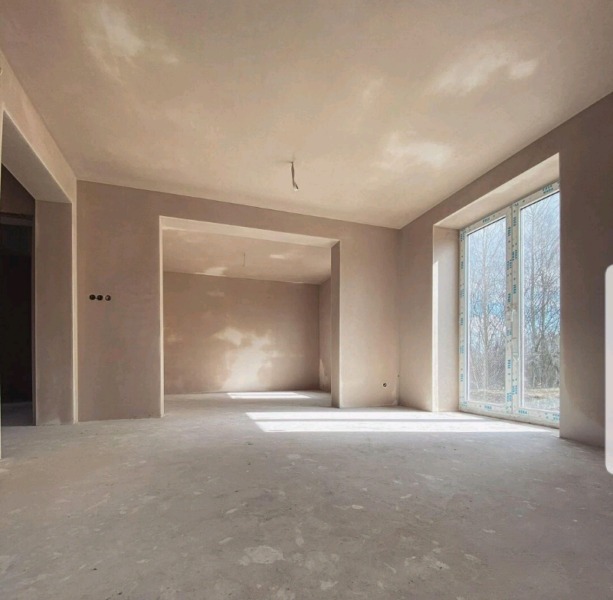 Wnętrze pomieszczenia w trakcie prac wykończeniowych, ściany i sufit pokryte świeżo nałożoną, jasną masą szpachlową, widoczne okno PCV i otwór drzwiowy.