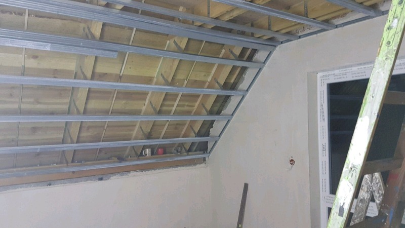 Stelaż z metalowych profili zamocowany do drewnianego stropu pod skosem, przygotowany pod montaż okładziny, widoczna ściana z oknem i drabina.