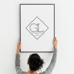 WisniewskiProduction - Osoba z tyłu trzyma czarną ramkę z minimalistycznym logo 'GL Galeria Lirka' na białym tle ściany.