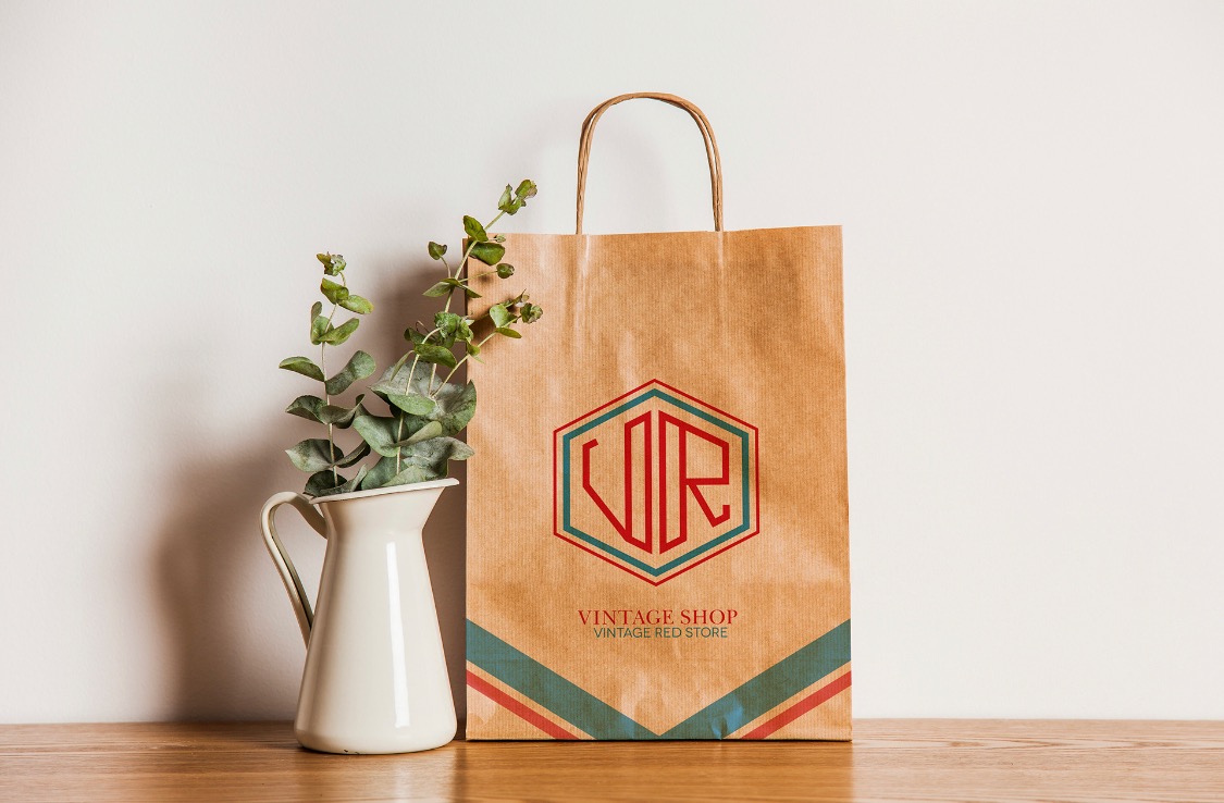 Brązowa papierowa torba z logo w kształcie sześciokąta, zawierającym litery VR, oraz napisem 'Vintage Shop Vintage Red Store', obok biały dzbanek z zielonymi gałązkami na drewnianym blacie.