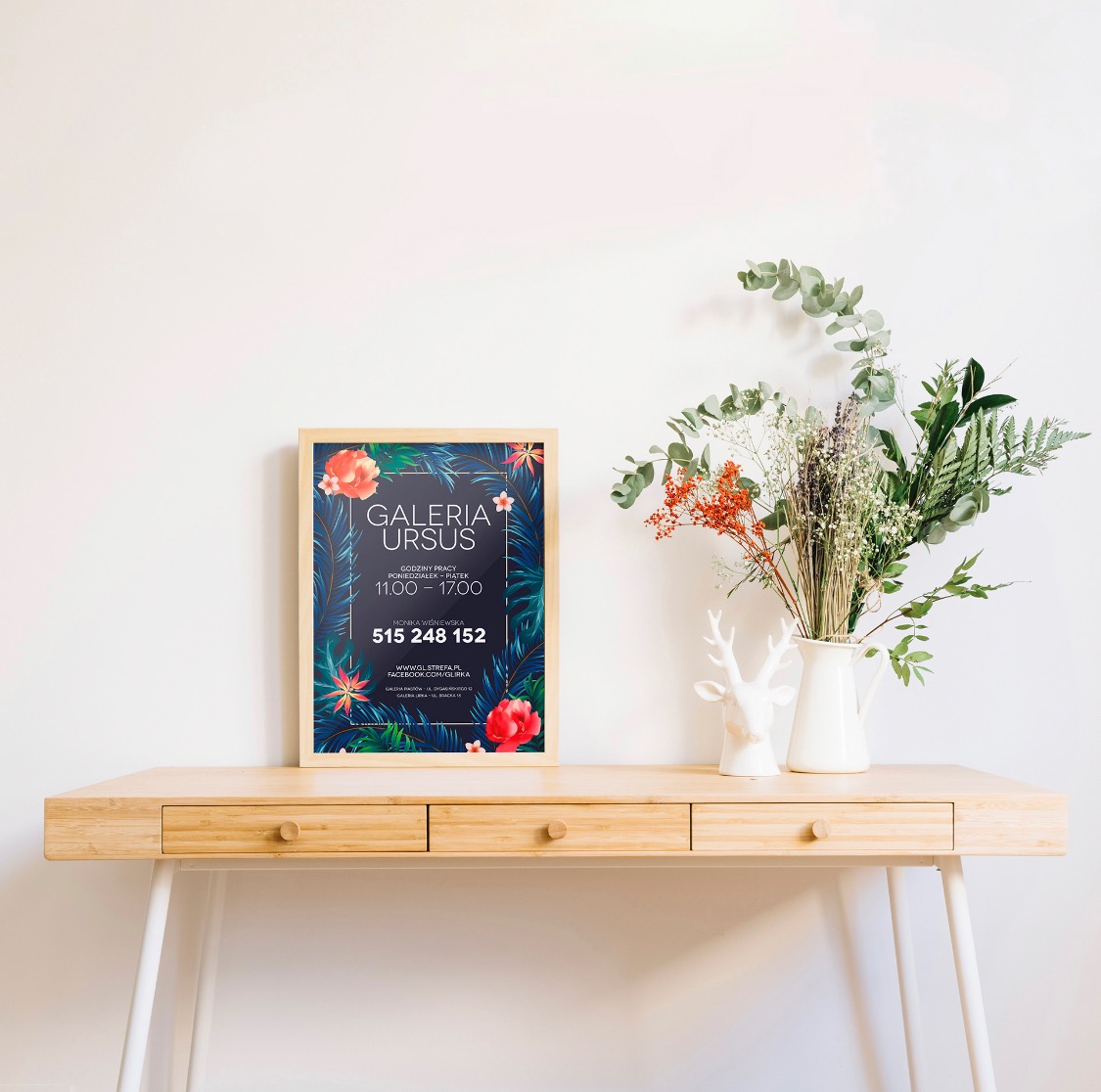 Plakat Galerii Ursus z motywem tropikalnym w ramce, stojący na drewnianej komodzie obok wazonu z kwiatami i figurki jelenia.