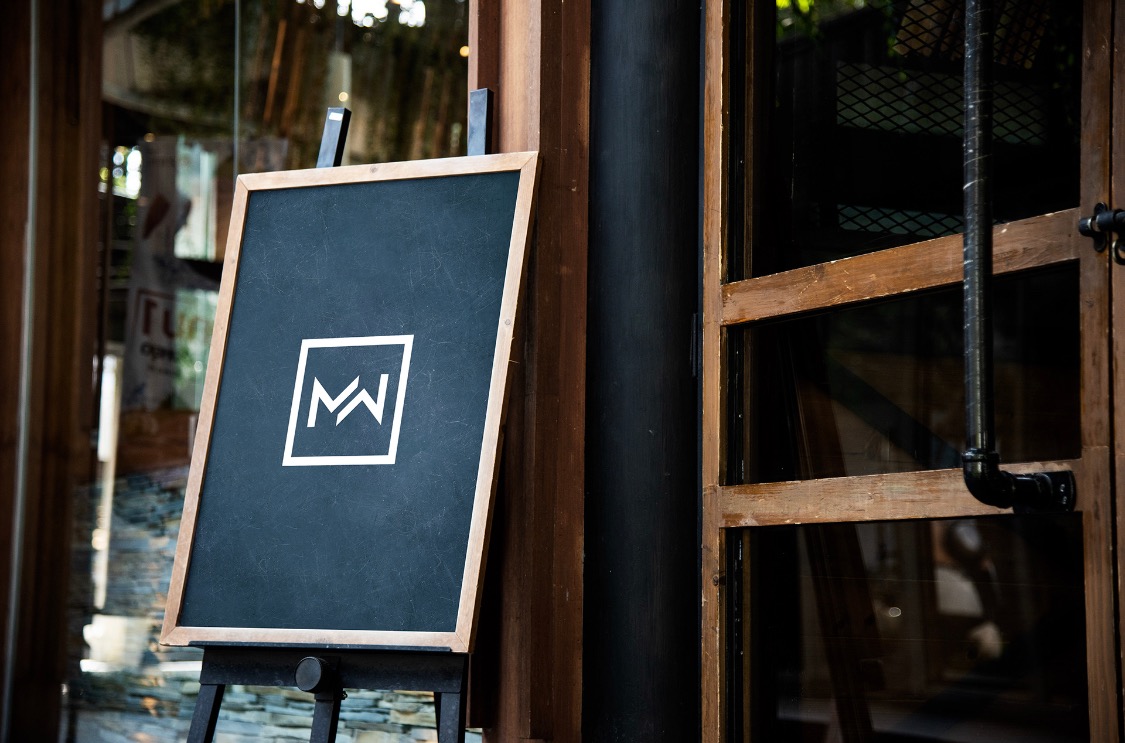 Tablica z logo MW na stojaku przed wejściem do budynku z drewnianymi drzwiami i oknami.