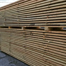 Stos jasnej tarcicy budowlanej ułożonej warstwami, z widocznymi przekładkami między niektórymi warstwami, na szarym podłożu.