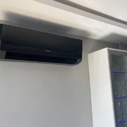 Czarna jednostka klimatyzacji marki Gree zamontowana na szarej ścianie w nowoczesnym wnętrzu, obok białej witryny z podświetleniem LED.