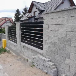 Fragment nowo wzniesionego ogrodzenia z szarego kamienia łupanego i czarnych poziomych paneli, widoczne ułożone obok kostki brukowe i ż&oacute;łta skrzynka gazowa, w tle dom z szarą dach&oacute;wką.