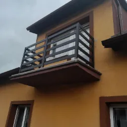 Czarna, metalowa balustrada balkonowa z poziomymi szczeblami na balkonie z betonową płytą, widok z dołu na żółtą elewację budynku z brązowymi obramowaniami okien i dachem w tle.